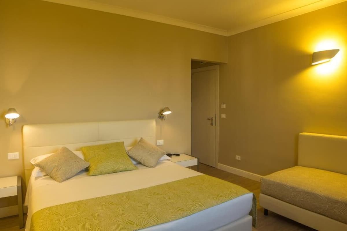 Una habitación de hotel con una cama, mesitas de noche, una silla y una puerta.