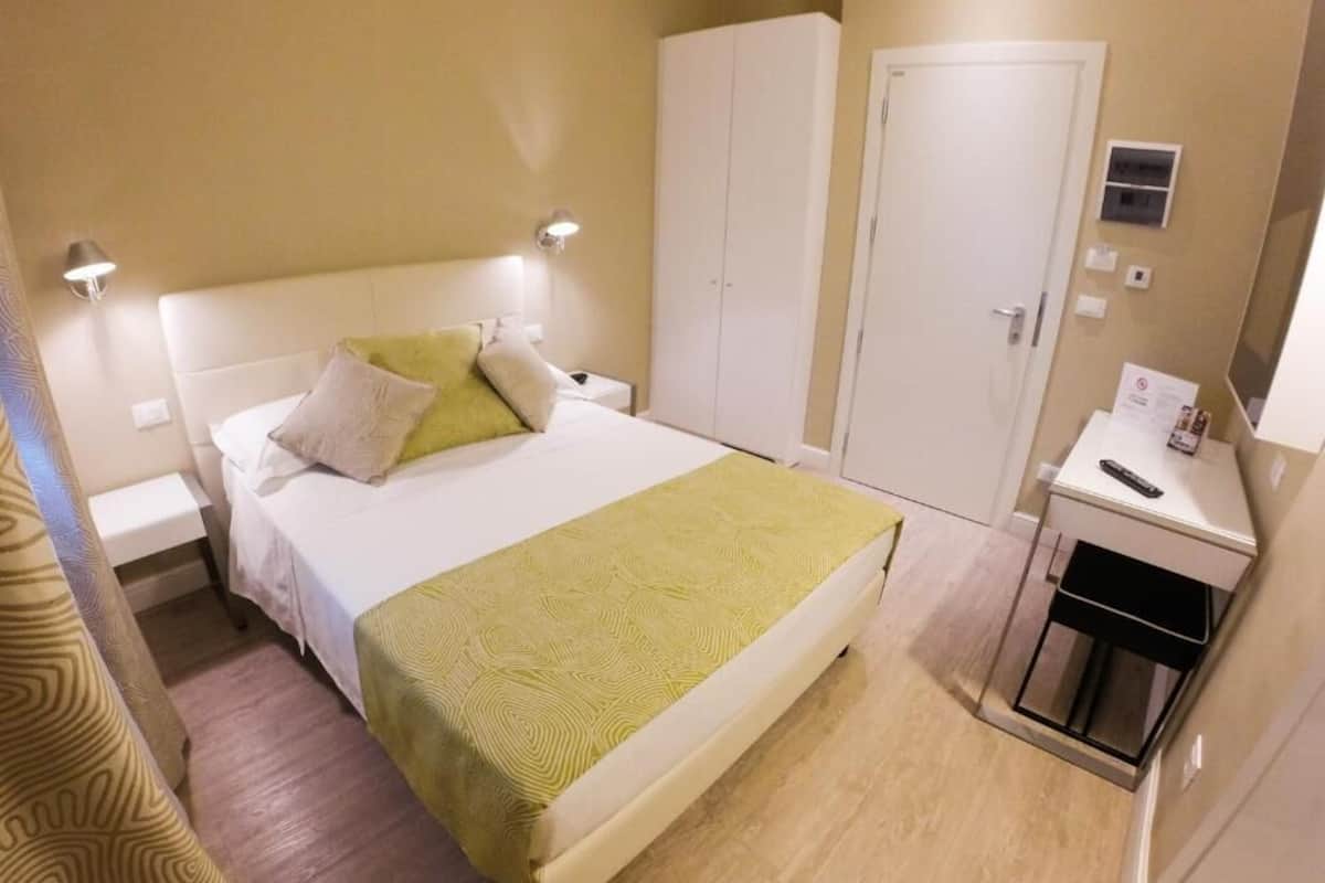 Una habitación de hotel con cama, mesitas de noche, armario, espejo y una pequeña kitchenette.