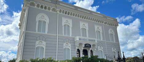 A large, historic building with a symmetrical facade, multiple windows with shutters, and a central entrance with a balcony.
