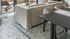 A modern living room with a sofa, a dining table, and a TV. There are large windows with a view of greenery outside. - Myrtia Beach House (Chania)