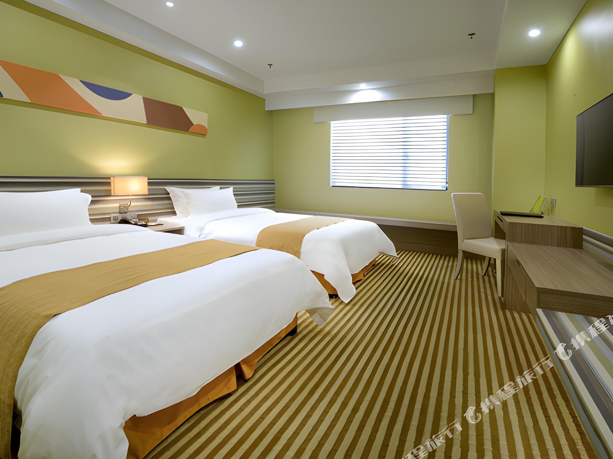 Ein Hotelzimmer mit zwei Betten, einem Schreibtisch, einem Stuhl, einem Fernseher und einem Fenster mit Jalousien.