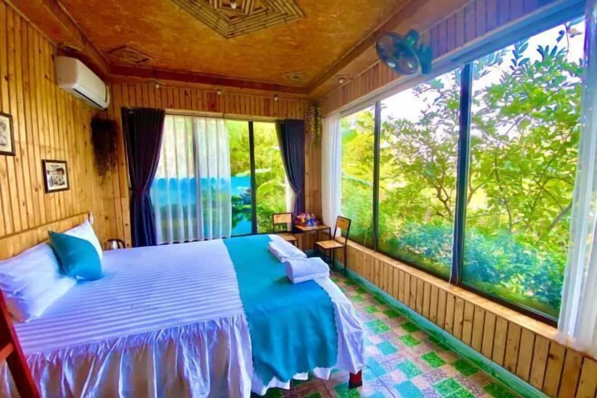 Quarto de cabana de madeira com cama, cortinas e vista para vegetação.