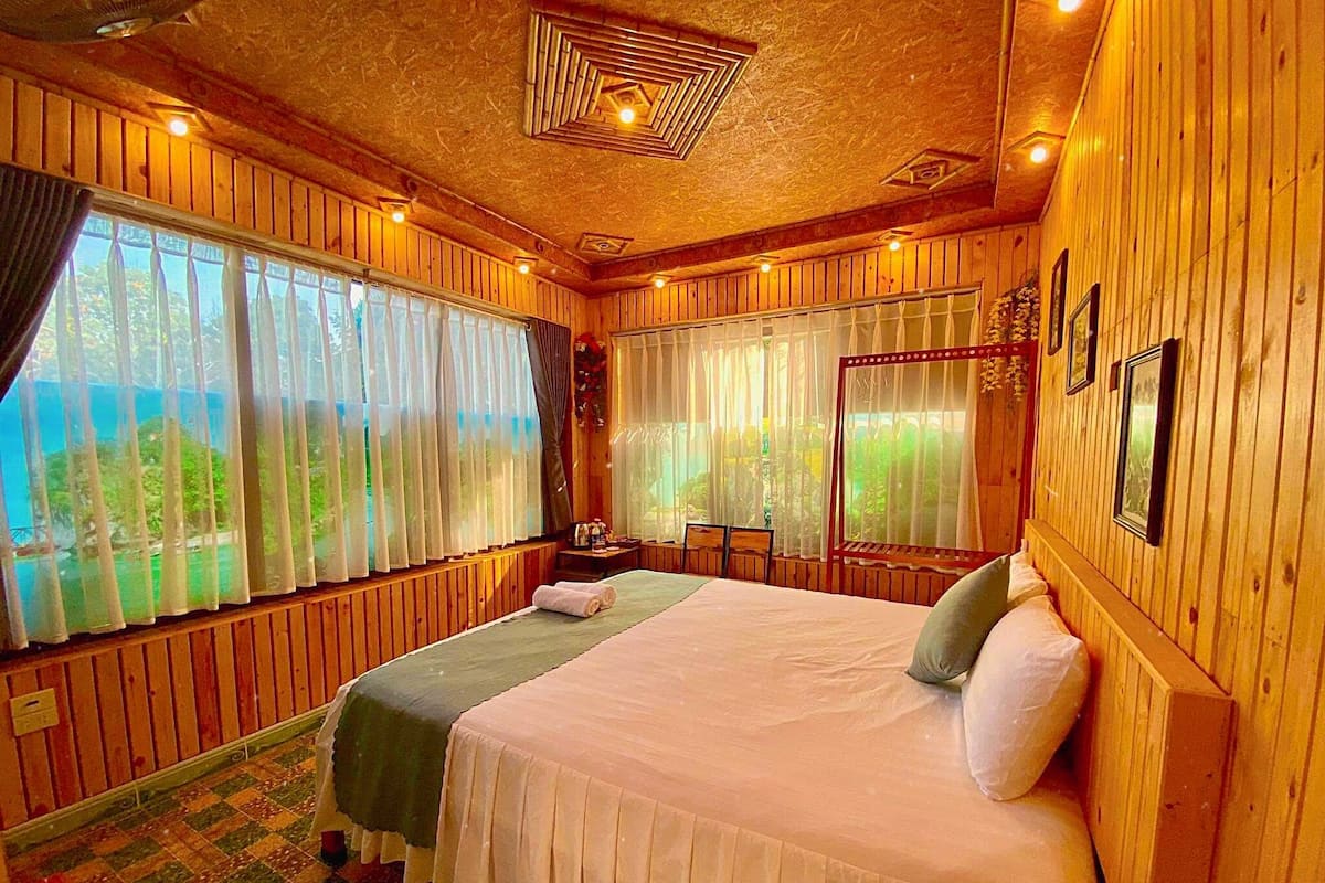 Um quarto de madeira com uma cama, uma cadeira e uma janela com cortinas.