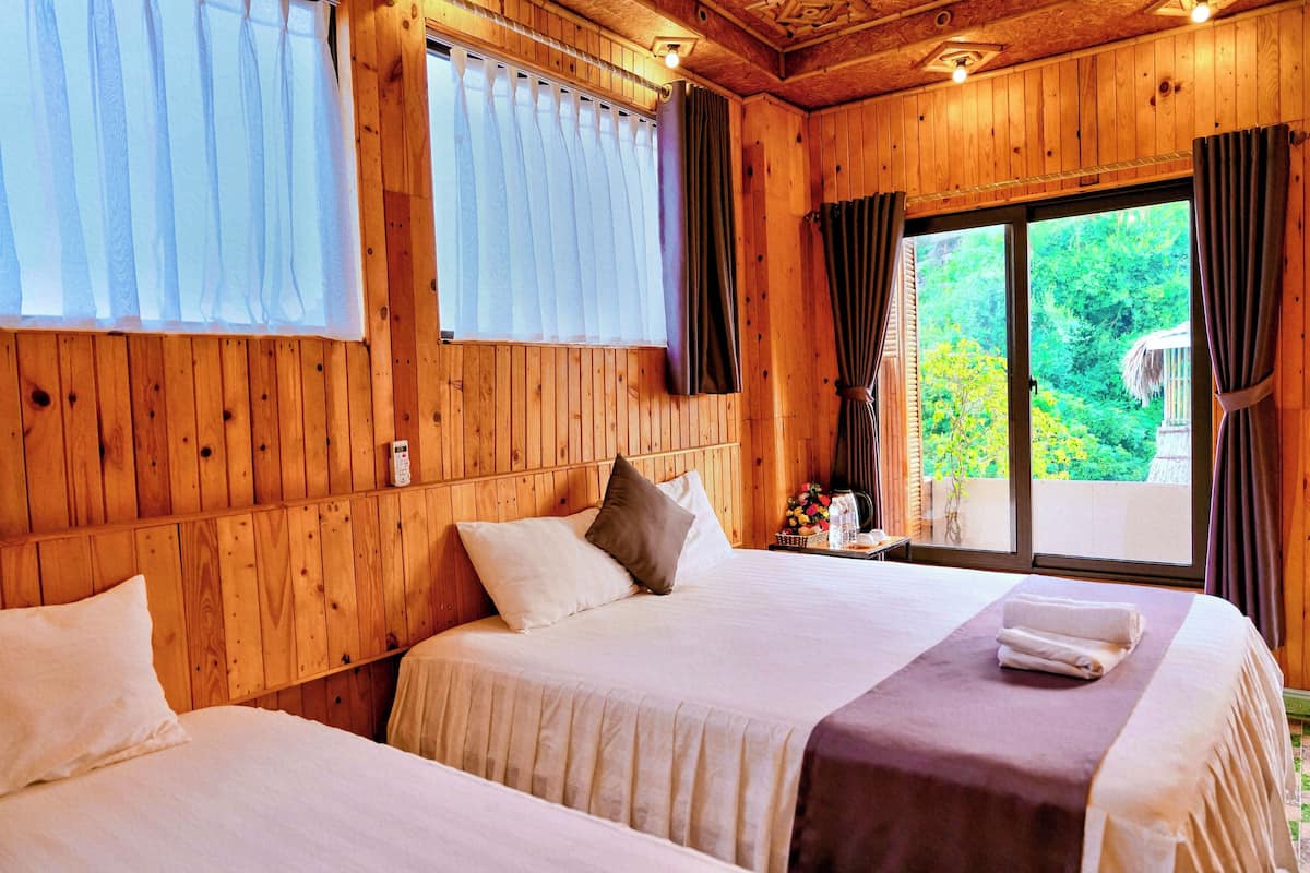 Quarto de cabana de madeira com duas camas, uma janela com cortinas e vista para o verde lá fora.