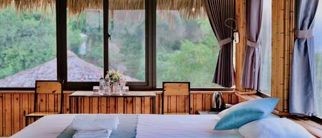 A room with a bed, a table with chairs, and large windows offering a view of greenery.