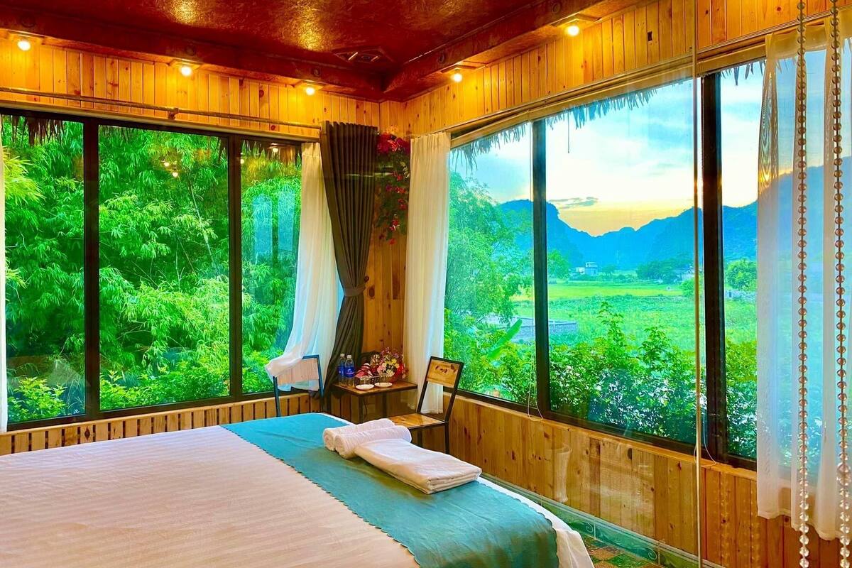 Quarto com uma cama grande, uma mesa pequena com uma cadeira e vista para uma paisagem verde através de grandes janelas.