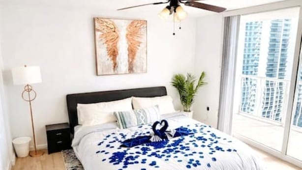 A bedroom with a bed, a ceiling fan, a large window, and a plant. - Brickell Bay 3505 (Miami)