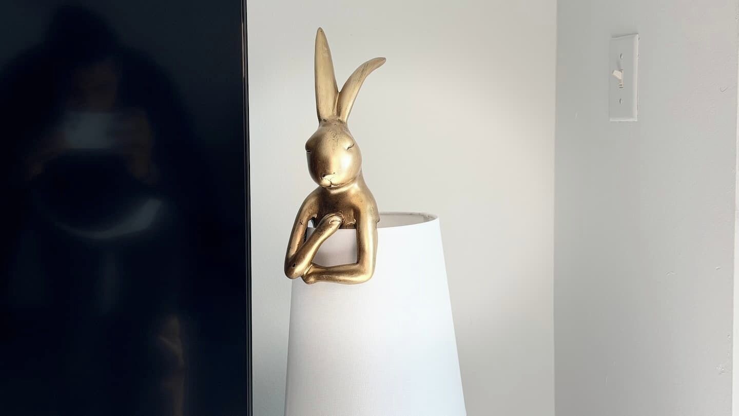 A gold rabbit sculpture on a white cylindrical object, possibly a lamp, in a room with a plain wall and a visible electrical outlet.