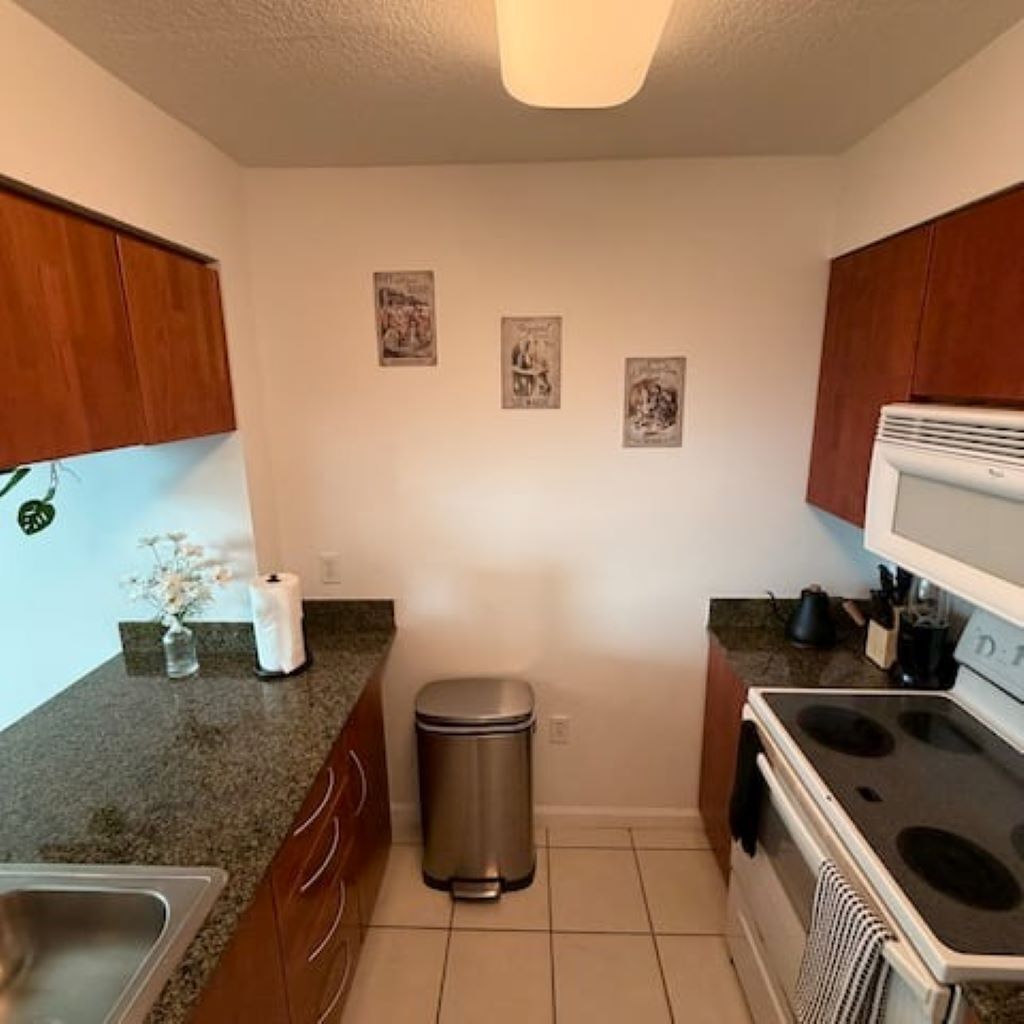 A compact kitchen with wooden cabinets, a stainless steel trash bin, a stove, and a countertop with a sink.