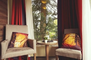 Deux fauteuils assortis avec des coussins identiques, une petite table ronde ornée d’une plante, et une vue sur les arbres à travers les rideaux.