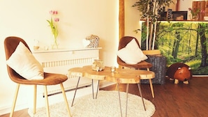 Un salon chaleureux avec une table ronde en bois, deux fauteuils marron avec des coussins blancs, un radiateur blanc, un tapis blanc, une étagère murale agrémentée d’objets décoratifs et un grand tableau sur le thème de la forêt.