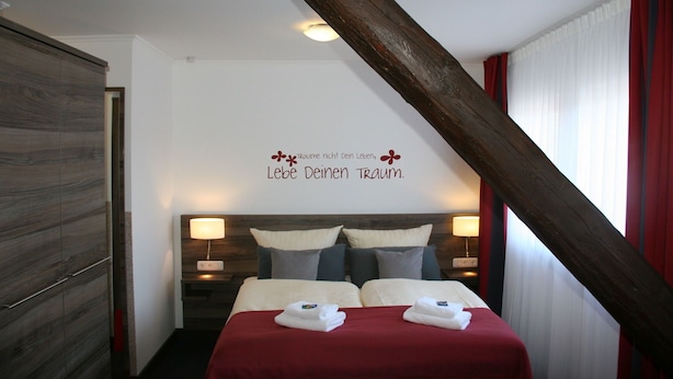 Une chambre d’hôtel avec une armoire en bois, un lit avec deux oreillers, une couverture rouge et un mur orné d’une citation inspirante.