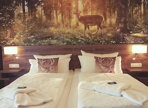 Un lit double avec deux oreillers, chacun orné d’un motif représentant un cerf, dans une chambre dont le mur est décoré d’une fresque sur le thème de la forêt.
