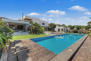 Un área de piscina moderna con una piscina azul cristalina, terraza de madera y una casa contemporánea con balcón.