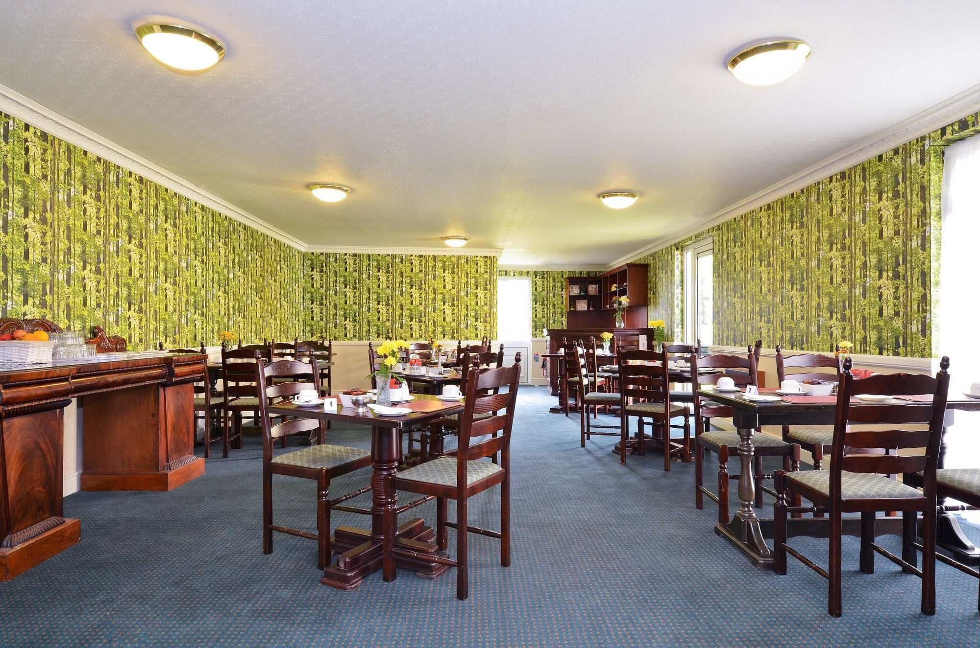 A dining area with wooden tables and chairs, green patterned wallpaper, and ceiling lights.
