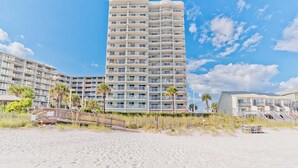 Un hotel frente a la playa con varios balcones, una playa de arena en primer plano y palmeras.