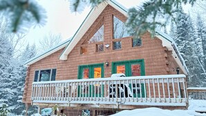 A wooden cabin with a balcony and green-framed windows, surrounded by snow-covered trees.