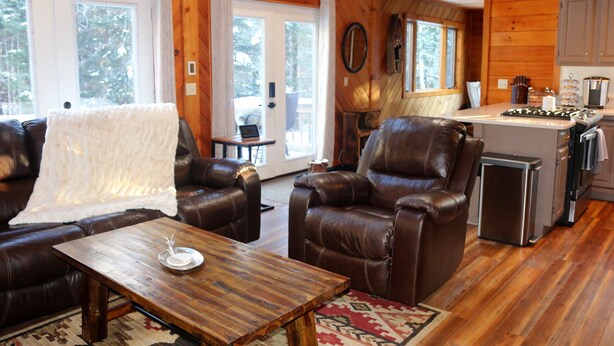 A cozy cabin interior with a living room featuring leather sofas and a wooden coffee table, a kitchen with modern appliances, and large windows offering a view of the surrounding forest.