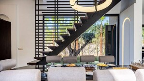Un área de comedor moderna con una escalera que lleva a un balcón, con un diseño minimalista, sillas tapizadas en gris y una mesa de vidrio.