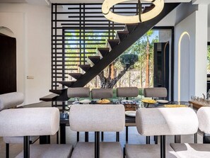 A modern dining area with a staircase leading to a balcony, featuring a minimalist design with grey upholstered chairs and a glass table. - Le Palais Zahira + Palais Mahora (2 villas) (Marrakech)