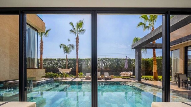 Un área de piscina moderna con paredes de vidrio transparente, sillones de descanso y palmeras.