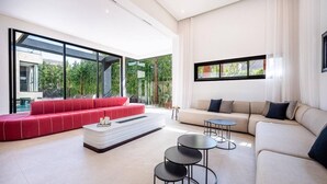 Una sala de estar moderna con un sofá seccional rojo, una mesa de centro blanca y un sofá color beige. Hay tres mesas redondas negras. Amplias ventanas ofrecen vistas a la vegetación exterior.