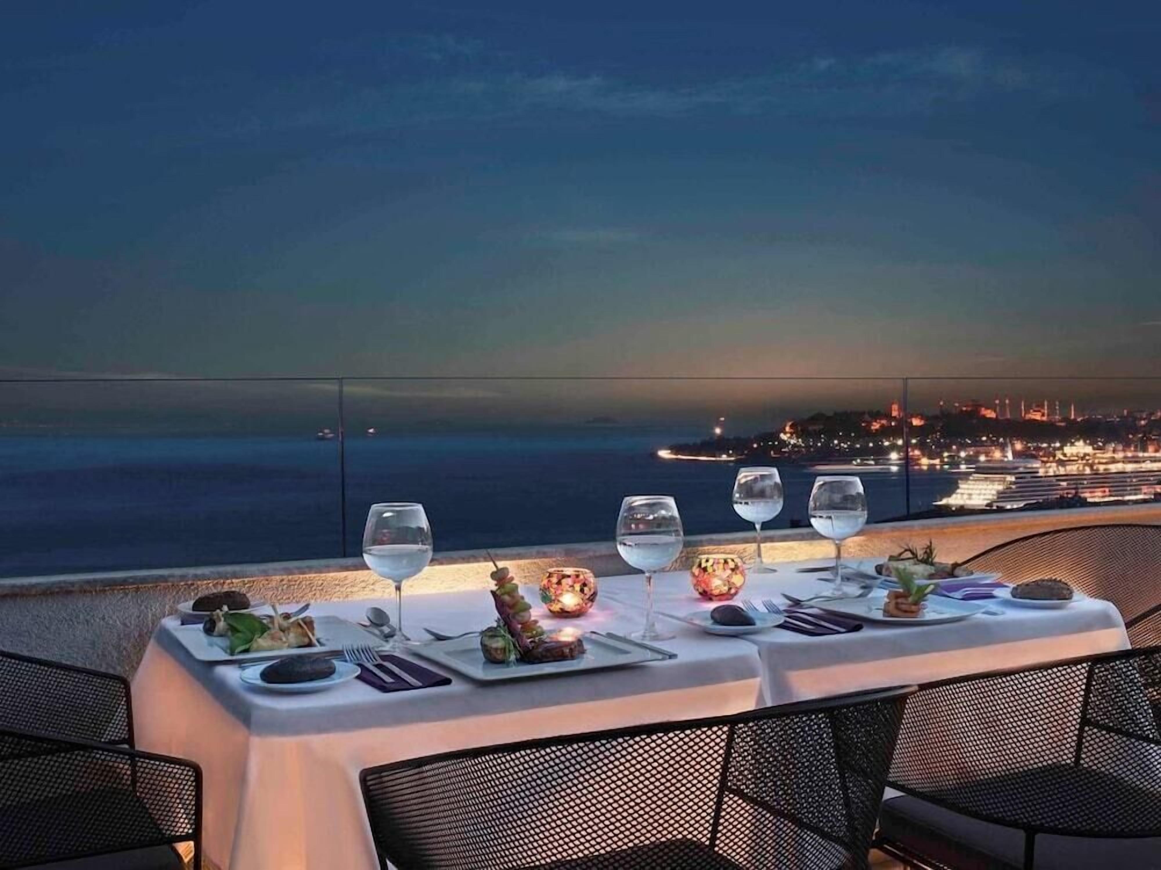 Una mesa dispuesta para una comida con vistas al mar y las luces de la ciudad al anochecer.
