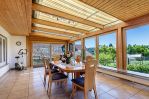 A spacious room with a wooden ceiling, large windows, a dining table with chairs, and an exercise bike. - Landzeit Apartment with Large Conservatory, Vineyard & Forest Views, 130 sqm (Efringen-Kirchen)