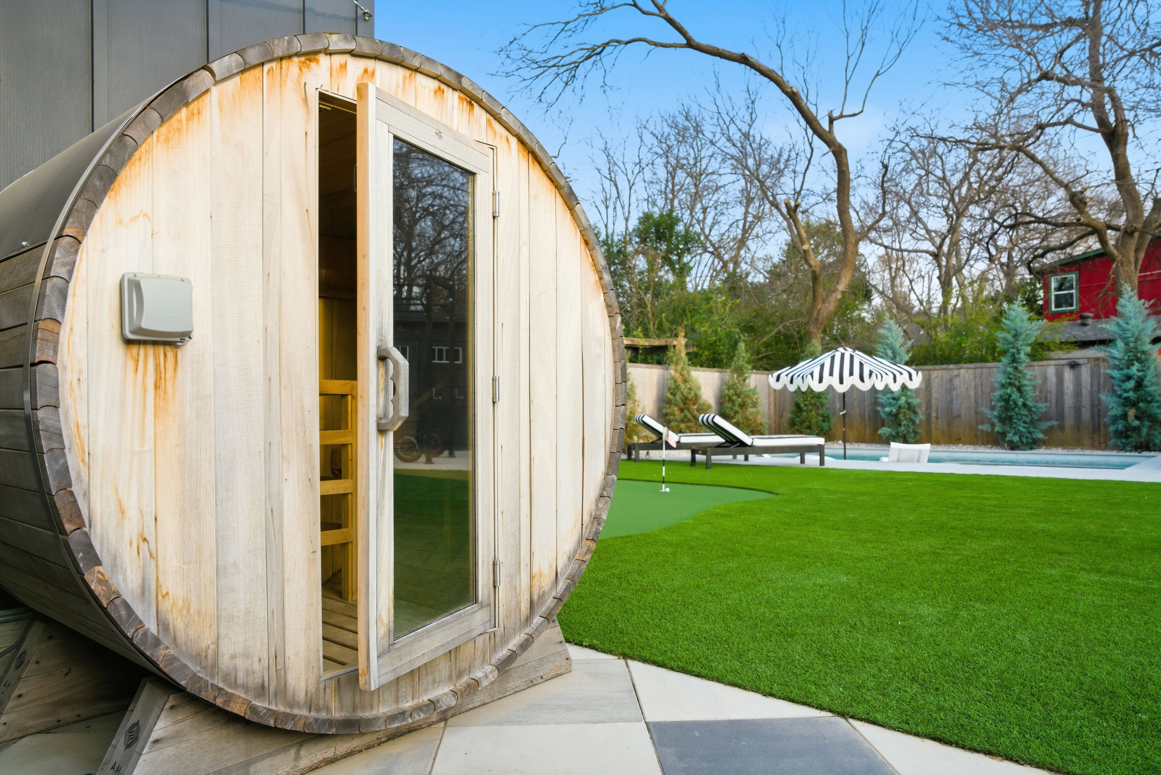 A wooden barrel sauna with a sliding glass door, a small patio area with a lawn, and a red building in the background.