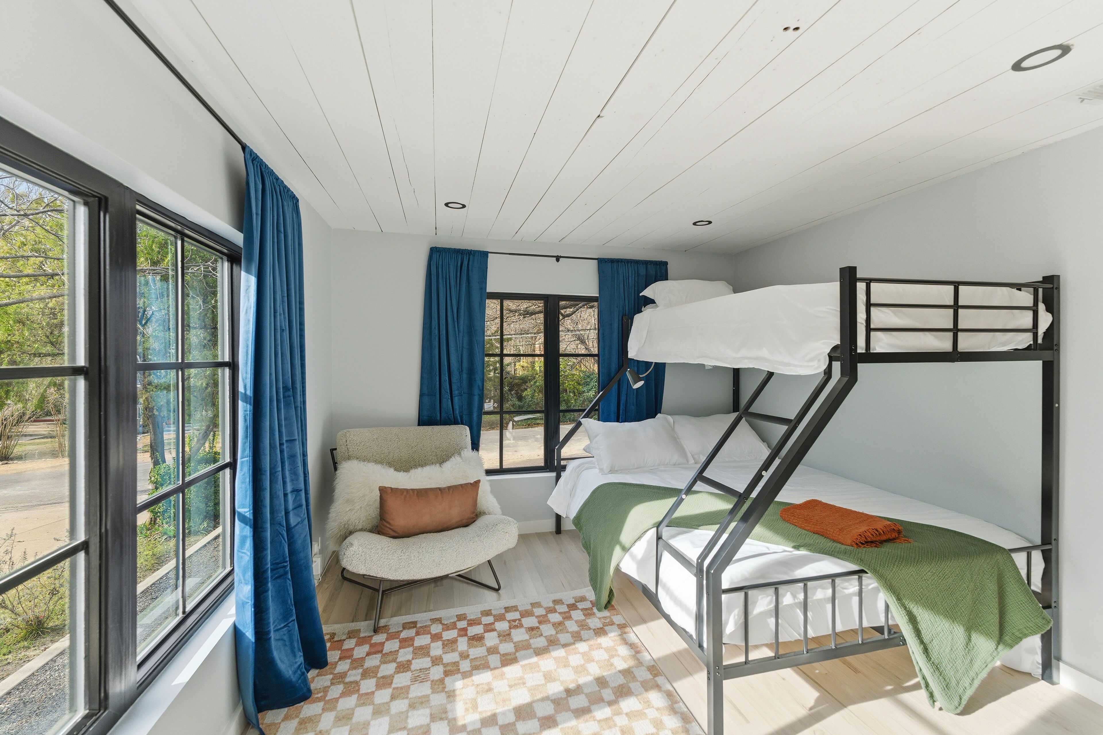 Une chambre avec des lits superposés, un tapis à carreaux, une chaise avec un coussin et une vue sur l’extérieur grâce à de grandes fenêtres.