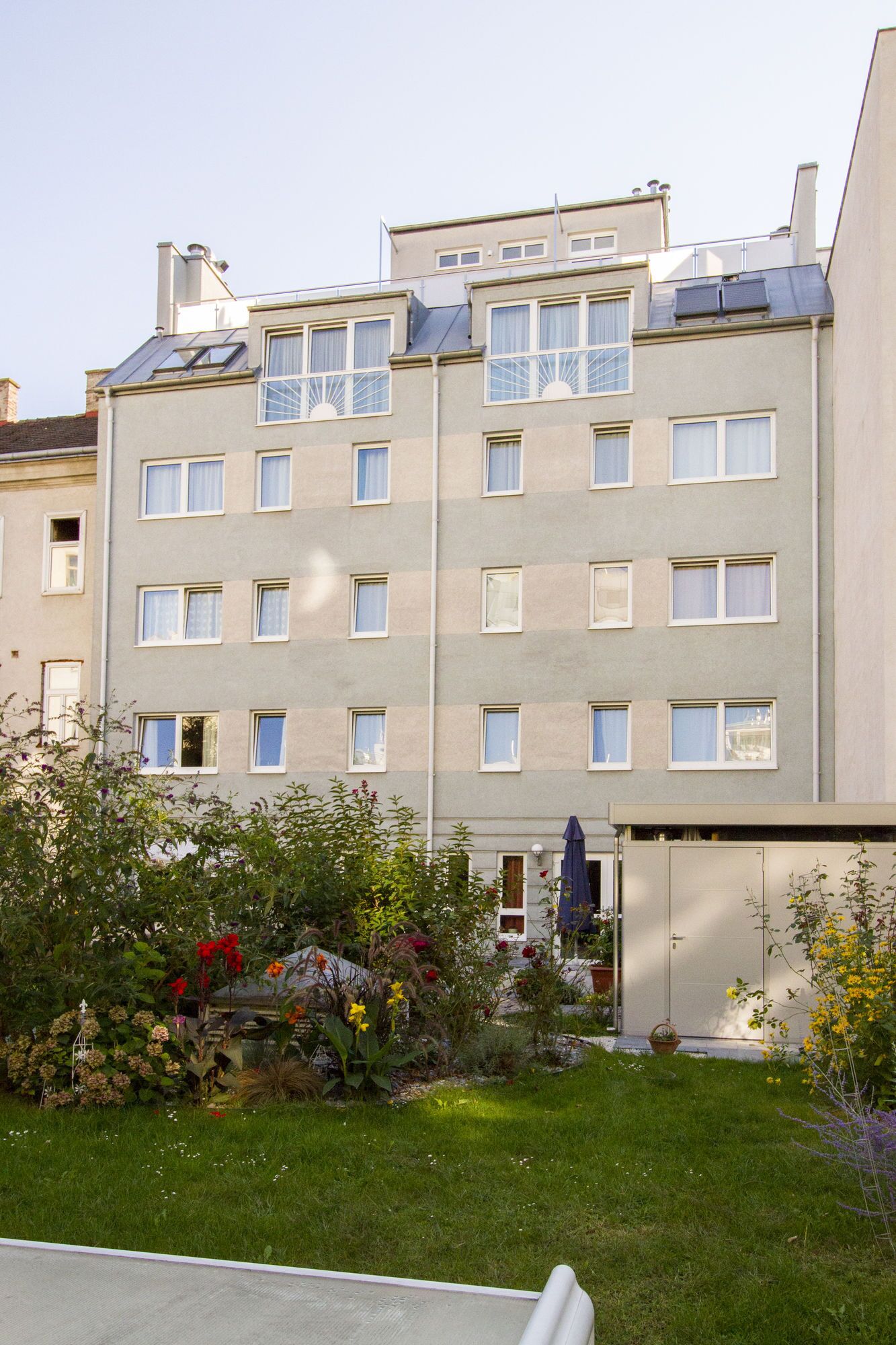 Un edificio residencial de varios pisos con techo plano, múltiples ventanas y un pequeño jardín con diversas plantas y flores.