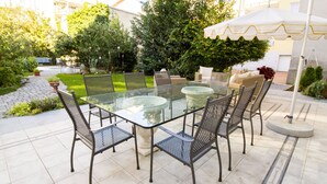 Eine Terrasse im Freien mit einem Glastisch und Metallstühlen, umgeben von viel Grün und einem weißen Sonnenschirm.