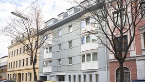 Un immeuble à plusieurs étages doté d’une façade moderne, plusieurs voitures garées et des arbres sans feuilles.