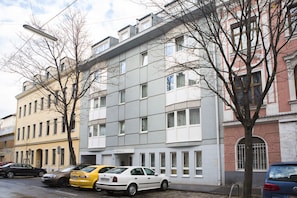 Un immeuble à plusieurs étages doté d’une façade moderne, plusieurs voitures garées et des arbres sans feuilles.