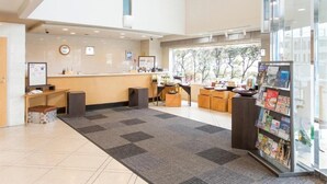 A reception area with a counter, seating, a magazine rack, and a view of trees through the window. - APA Hotel Shonan Tsujido Ekimae (Fujisawa)