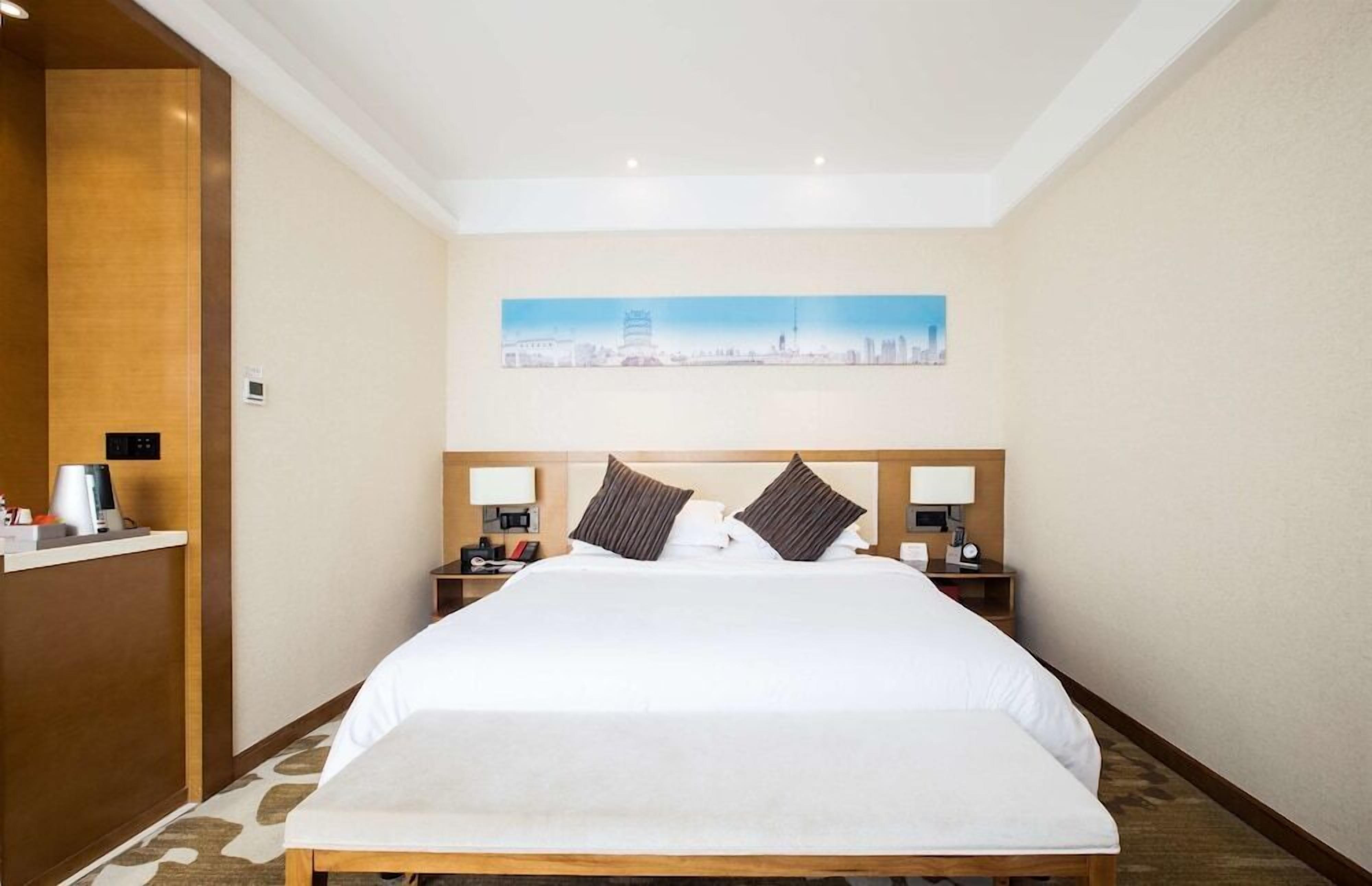 A hotel room with a large bed, two bedside tables with lamps, a cityscape painting on the wall, and a wooden panel on the left side.