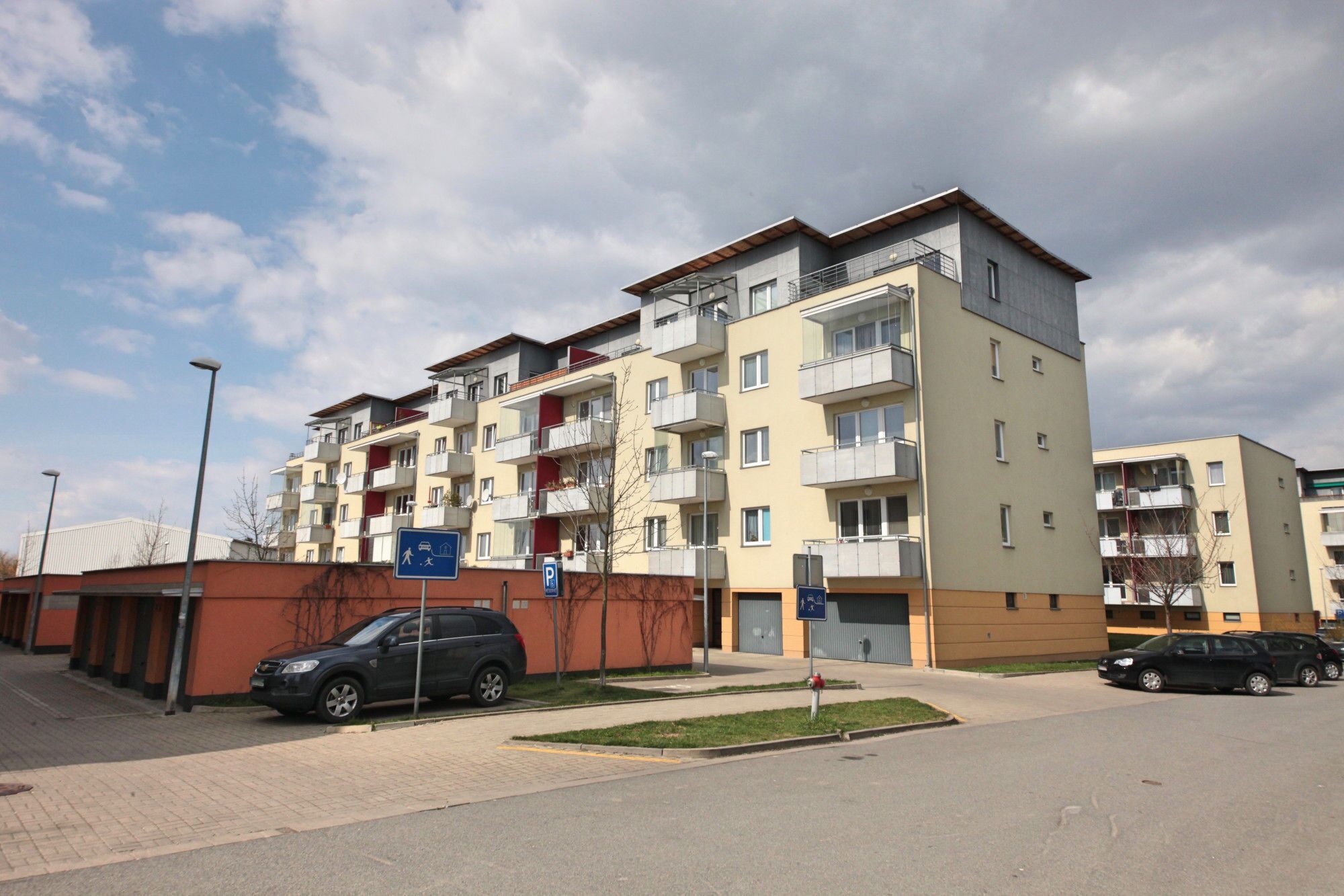 A multi-story residential building with balconies, a parking area, and a clear sky.