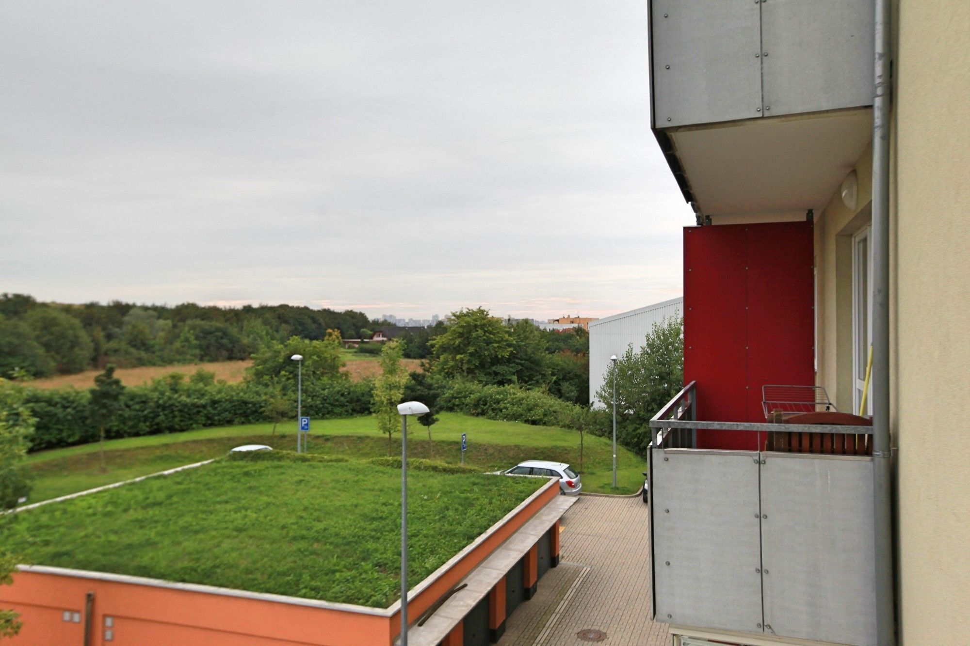 A view from a balcony overlooking a green lawn, a parking area, and a distant cityscape.