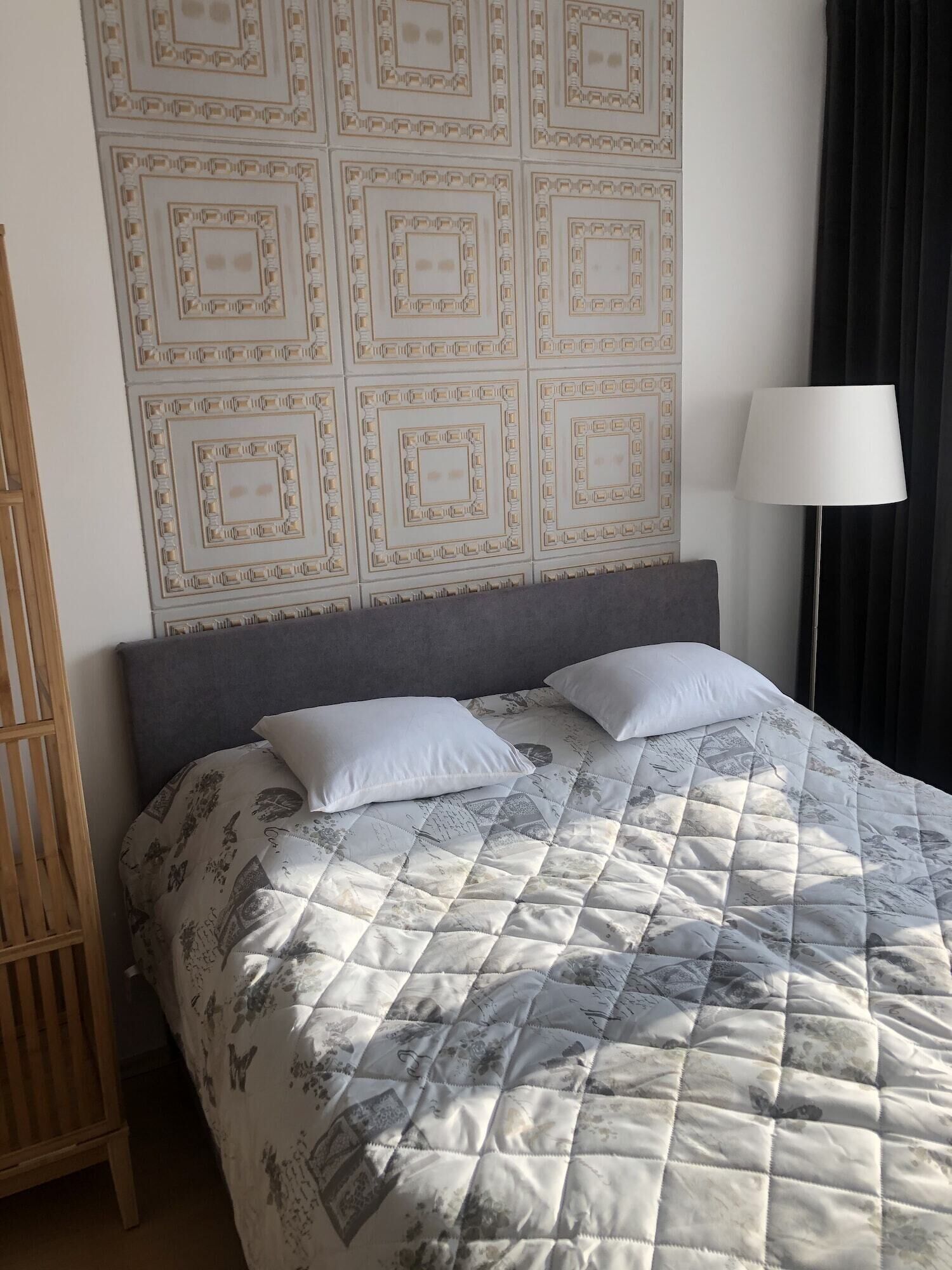 Cama bem arrumada com edredom acolchoado e duas almofadas, luminária de cabeceira e parede com padrão geométrico.