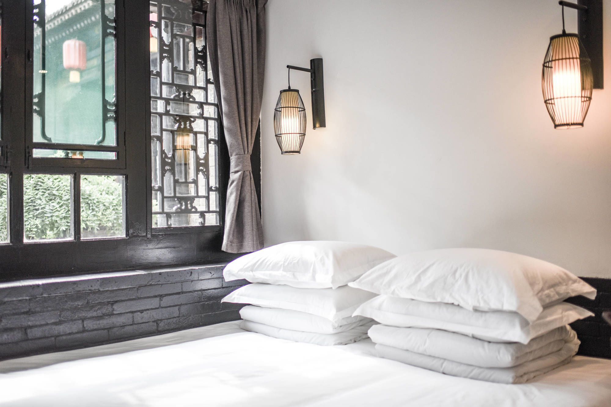 A neatly made bed with white linens, a window with decorative iron bars, and two wall-mounted light fixtures.