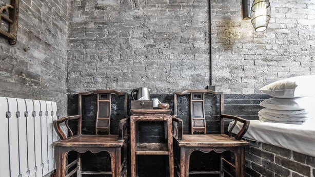 Een rustieke kamer met bakstenen muren, een metalen radiator en drie houten stoelen met metalen poten.