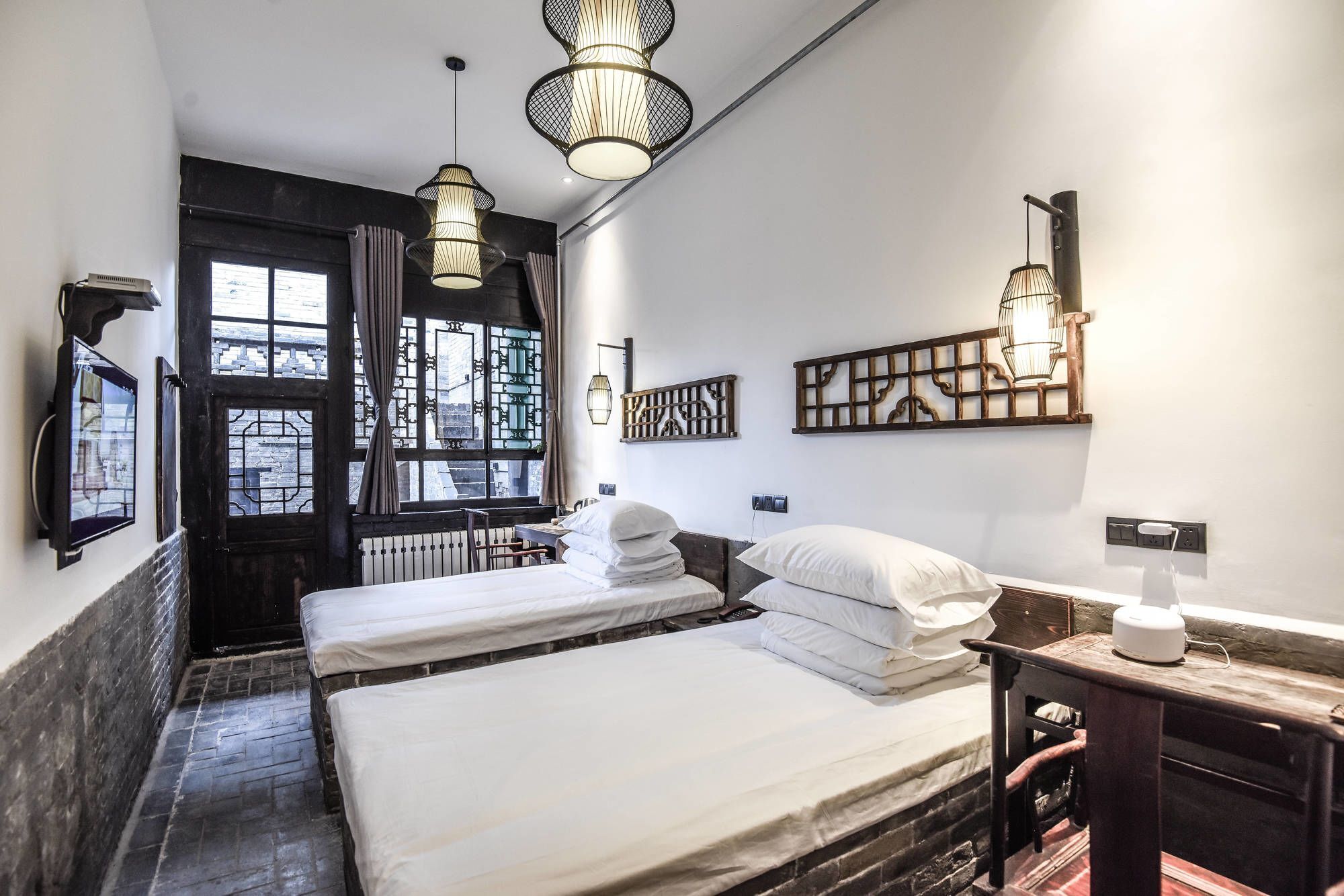 A hotel room with two beds, a window with curtains, a wall-mounted light fixture, and a small table with a kettle.