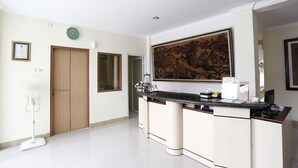 Ein Hotel-Empfangsbereich mit einer Holztür, einem großen traditionellen Gemälde, einer Rezeption mit Computer und einem Standventilator.