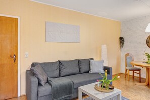 Uma sala de estar com um sofá cinzento, uma mesa de centro branca, uma mesa de jantar com cadeiras e uma obra de arte abstrata fixada na parede.