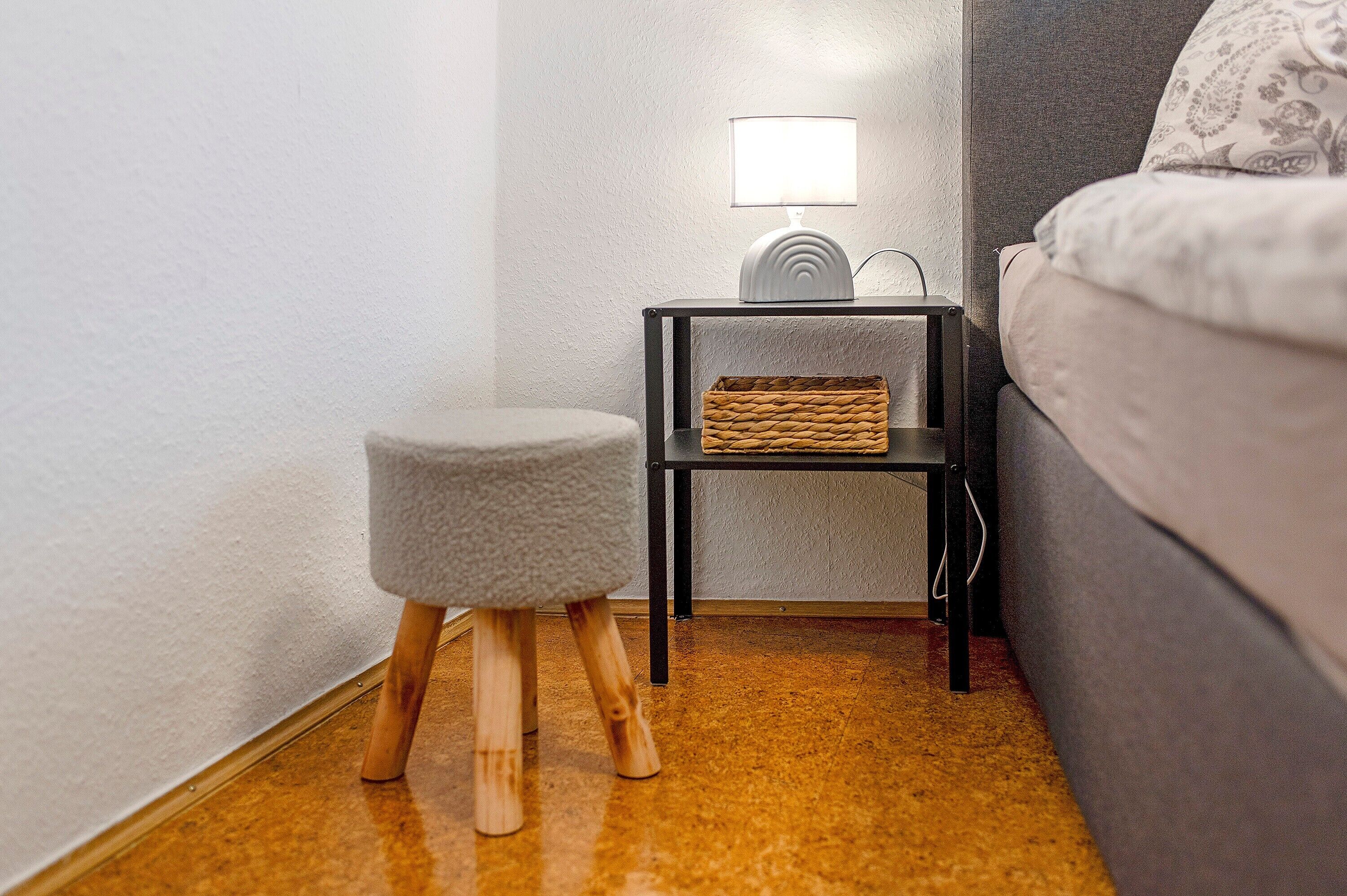 A corner of a room with a grey sofa, a small round stool, a black nightstand with a lamp, and a woven basket.