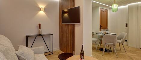 A modern living room with a sofa, a small round table with a bottle and glasses, a TV mounted on a wooden panel, and a dining area in the background.