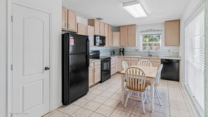 Une cuisine compacte avec un réfrigérateur noir, un four à micro-ondes et une cuisinière. On y trouve une table à manger avec quatre chaises. La cuisine est dotée de meubles de couleur claire et d’une fenêtre avec des stores.