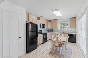 A compact kitchen with a black refrigerator, a microwave, and a stove. There is a dining table with four chairs. The kitchen has light-colored cabinets and a window with blinds. - Townhouse style condo 1 block from beach! (Indian Rocks Beach)