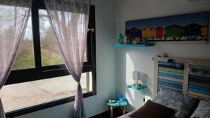 Une chambre avec un lit, une fenêtre ornée de rideaux légers, une petite table avec une lampe et une étagère fixée au mur, sur laquelle sont disposés des objets colorés.