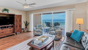A spacious living area with a large TV, wooden entertainment unit, and a balcony with a view of the ocean. - Casa de Playa 206 - Beach Front Condo (Indian Rocks Beach)